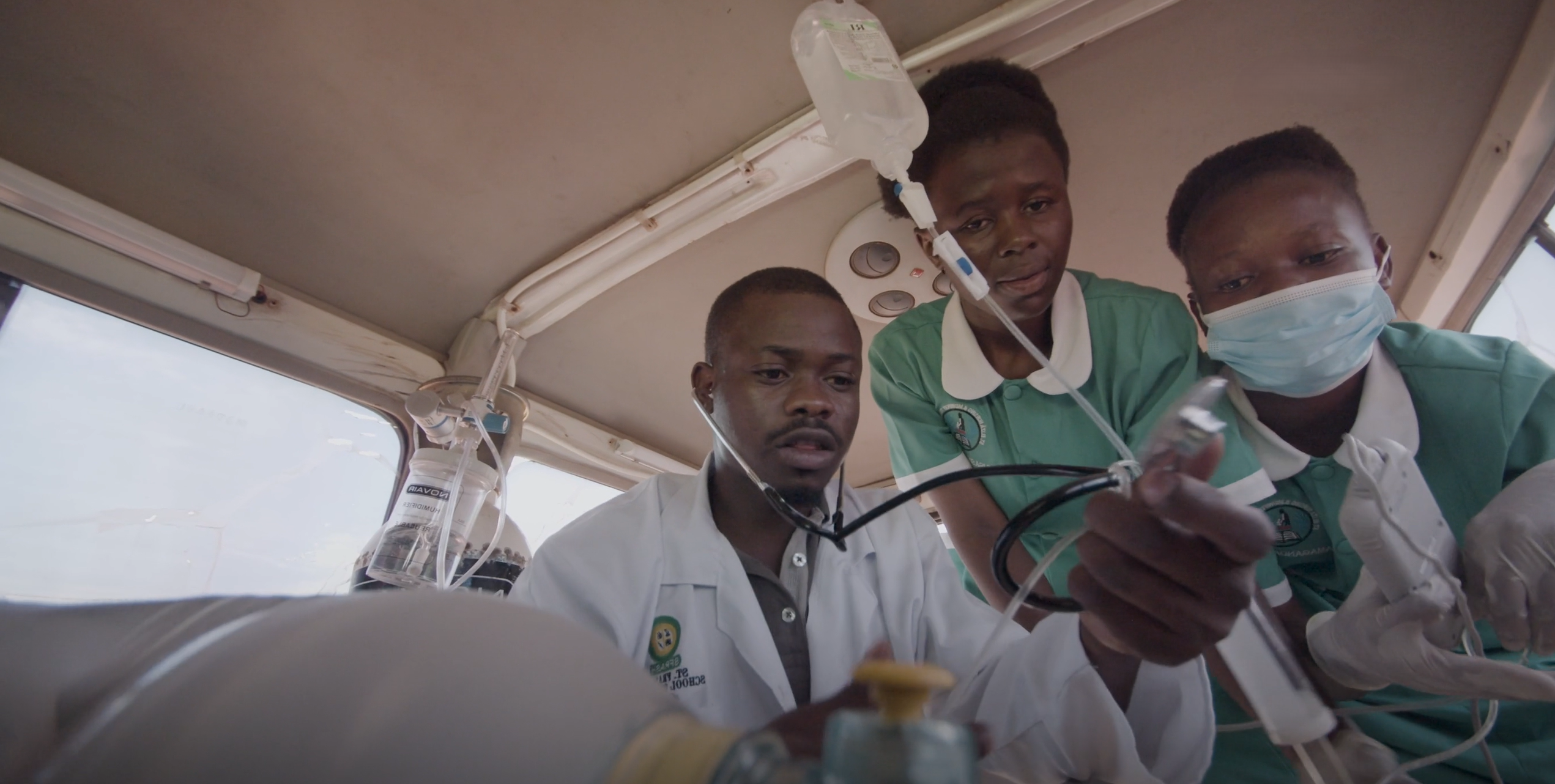 Inside an ambulance, a male healthcare worker uses a stethoscope to examine a patient's chest, assisted by two female healthcare workers in uniform holding medical equipment.
