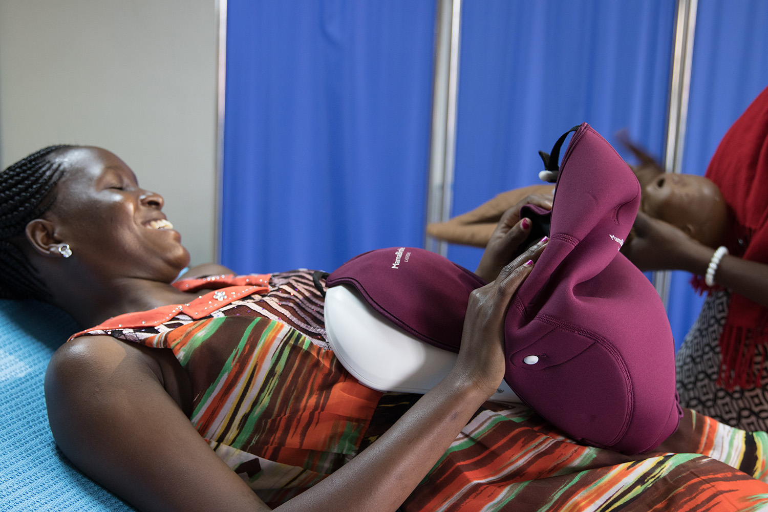 A smiling woman lying on a clinic bed with a MamaNatalie maternal simulator during a hands-on simulation training session.