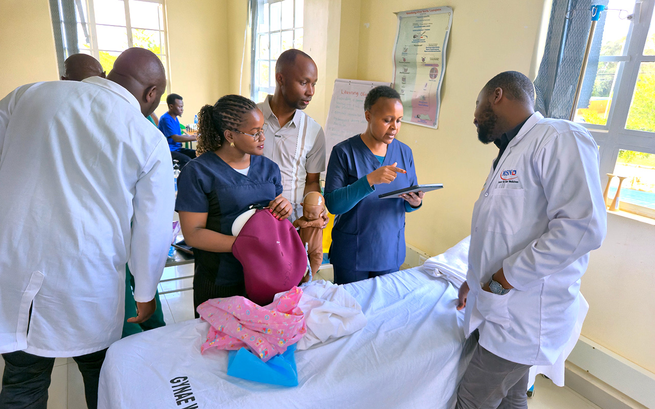 A close-up of a group of five people, including doctors and nurses, around a medical training table and looking down as a woman in blue scrubs holds and explains to them about a MamaNatalie simulator and a newborn simulator. In the background on the wall,